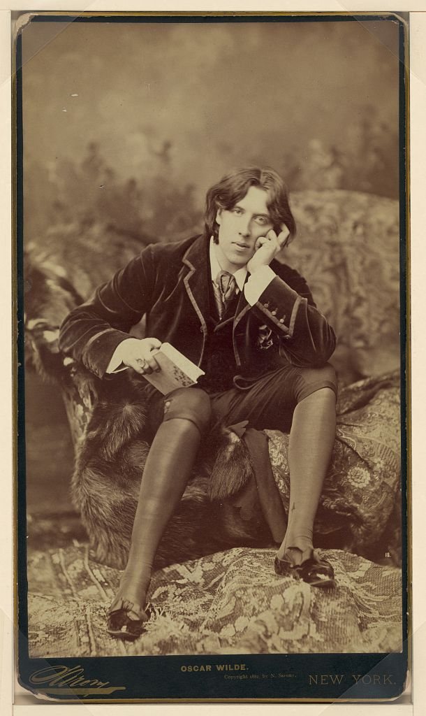 Sepia cabinet-card portrait of Oscar Wilde seated on a fur-draped divan, holding a book, chin resting on his hand.