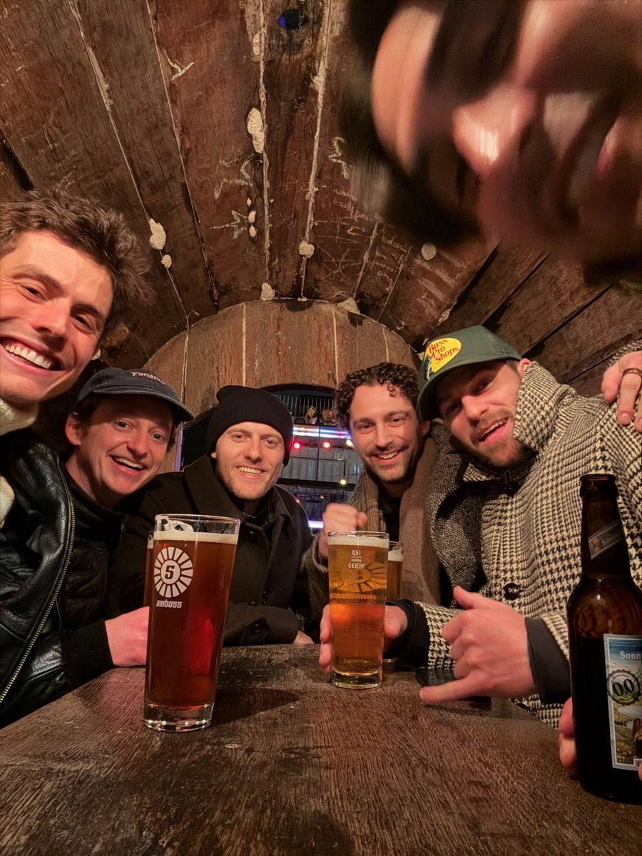 Five founders laughing around a table inside a wooden barrel booth, with beers, a hand blurring the corner of the frame.